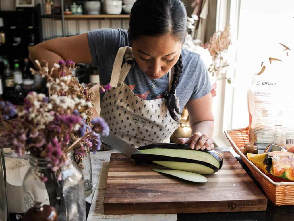 A woman in a blue shirt and apron is preparing food in a kitchen. She is carefully slicing a large eggplant on a wooden cutting board. The kitchen is adorned with dried flowers and various cooking ingredients.